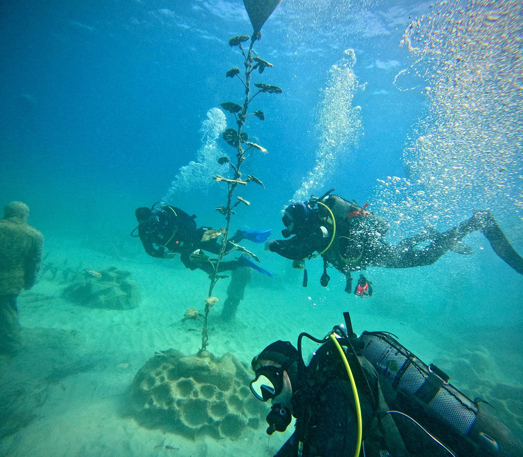 Three RAF Brize Norton Sub Aqua Club scuba divers exploring a shallow underwater park with sandy seabed, rising air bubbles, and structural marine installations.